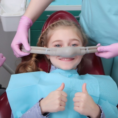 Young girl giving thumbs up in dental chair with nitrous oxide mask over nose