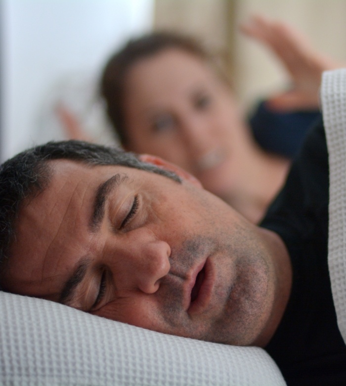 Man snoring while woman in bed next to him holds up her hands in frustration